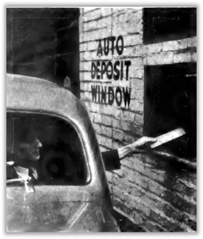 A black and white photo shows a automobile driver passing a piece of paper into a window labeled AUTO DEPOSIT WINDOW at the side of a brick building.