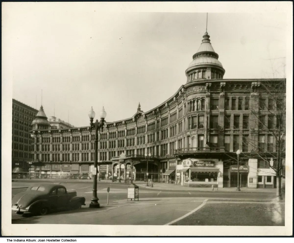A black and white photo of the 4-story English Hotel, forming a semi-circle around Monument Circle. On the corner of the building is a taller tower.