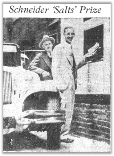 A black and white photo titled Schneider ‘Salts’ Prize shows Louis Schneider and another man standing beside a window at the side of a brick window, handing papers into the window. A third man in the driver seat of an automobile tilts his head out the car window.